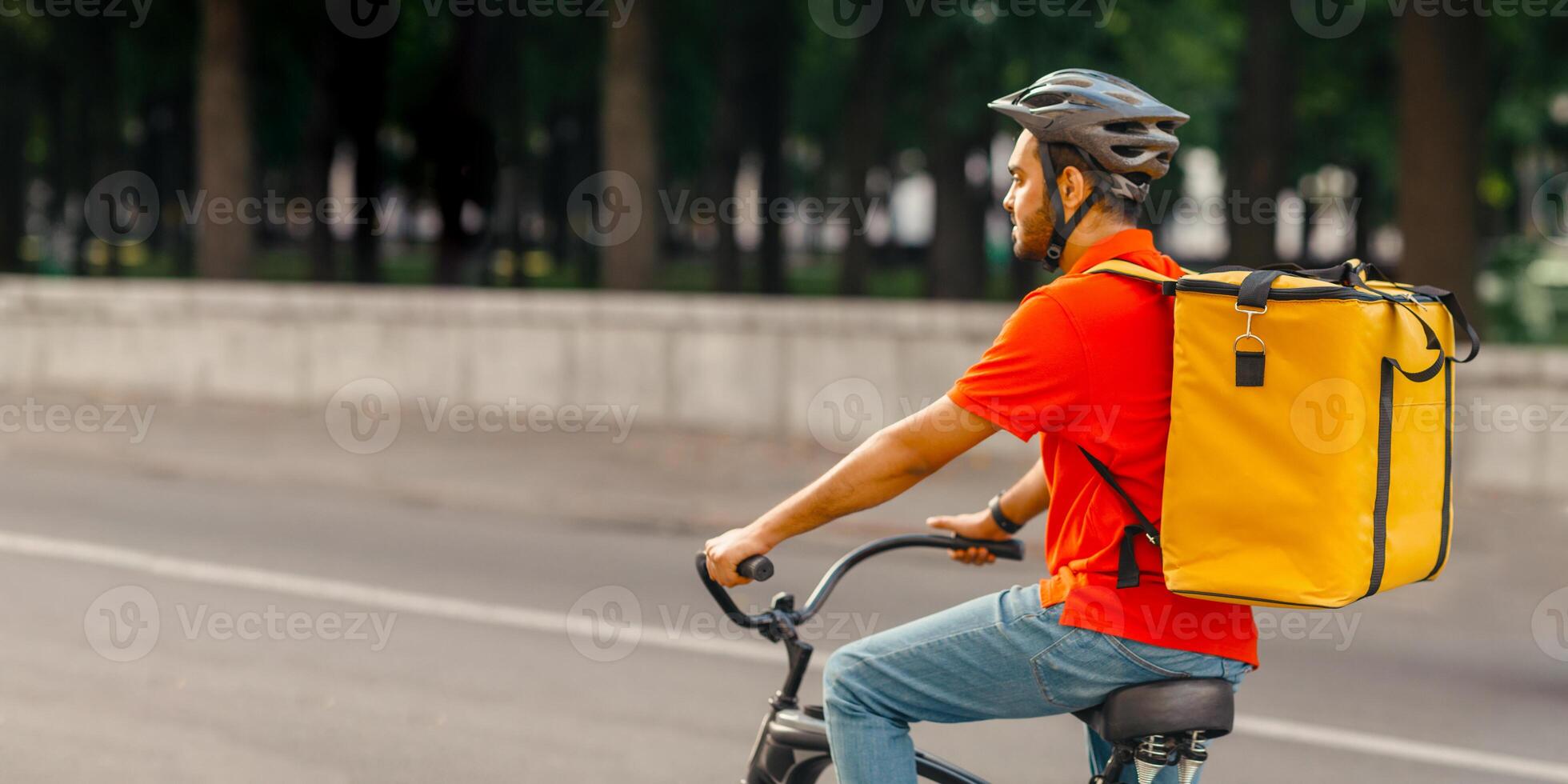 Eco quick delivery, city food gear service and online ordering. Millennial indian man with beard in uniform, protective helmet and yellow bag rides on bicycle on road, outside, side view, empty space photo
