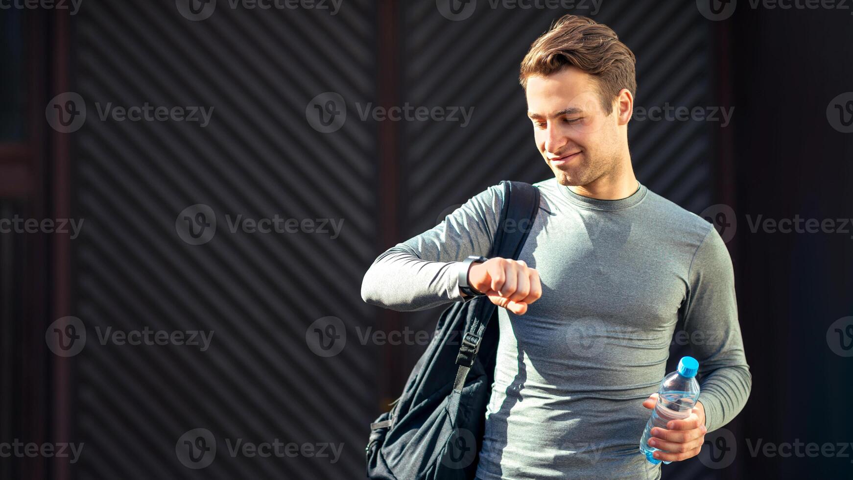 Athletic style and preparation for run. Young smiling man goes to training with sports bag and in sportswear, looks at tracker on hand and holds water photo