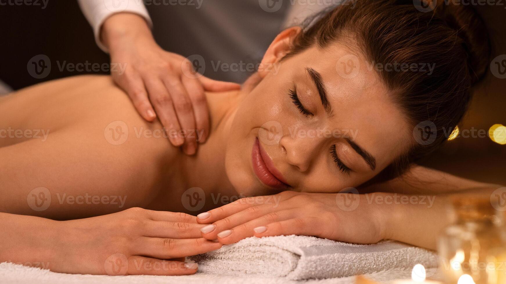Joyful young woman having healing body massage with aromatherapy at luxury spa decorated with lightened candles. Female therapist rubbing sleeping lady shoulders, giving her relaxing massage, closeup photo