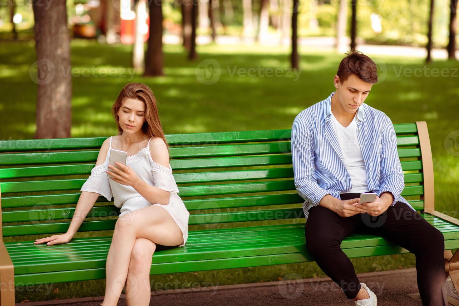 Gadget addiction and relationship problems. Millennial couple with smartphones sitting on bench at park, ignoring each other photo