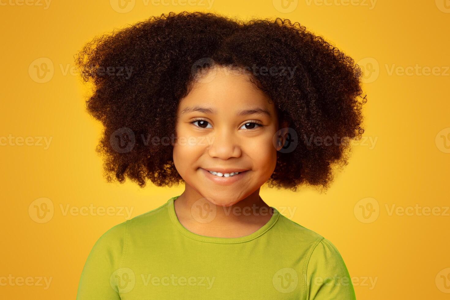 Portrait Of Adorable Curly Little Black Girl Smiling At Camera While Standing Over Yellow Background In Studio, Closeup photo