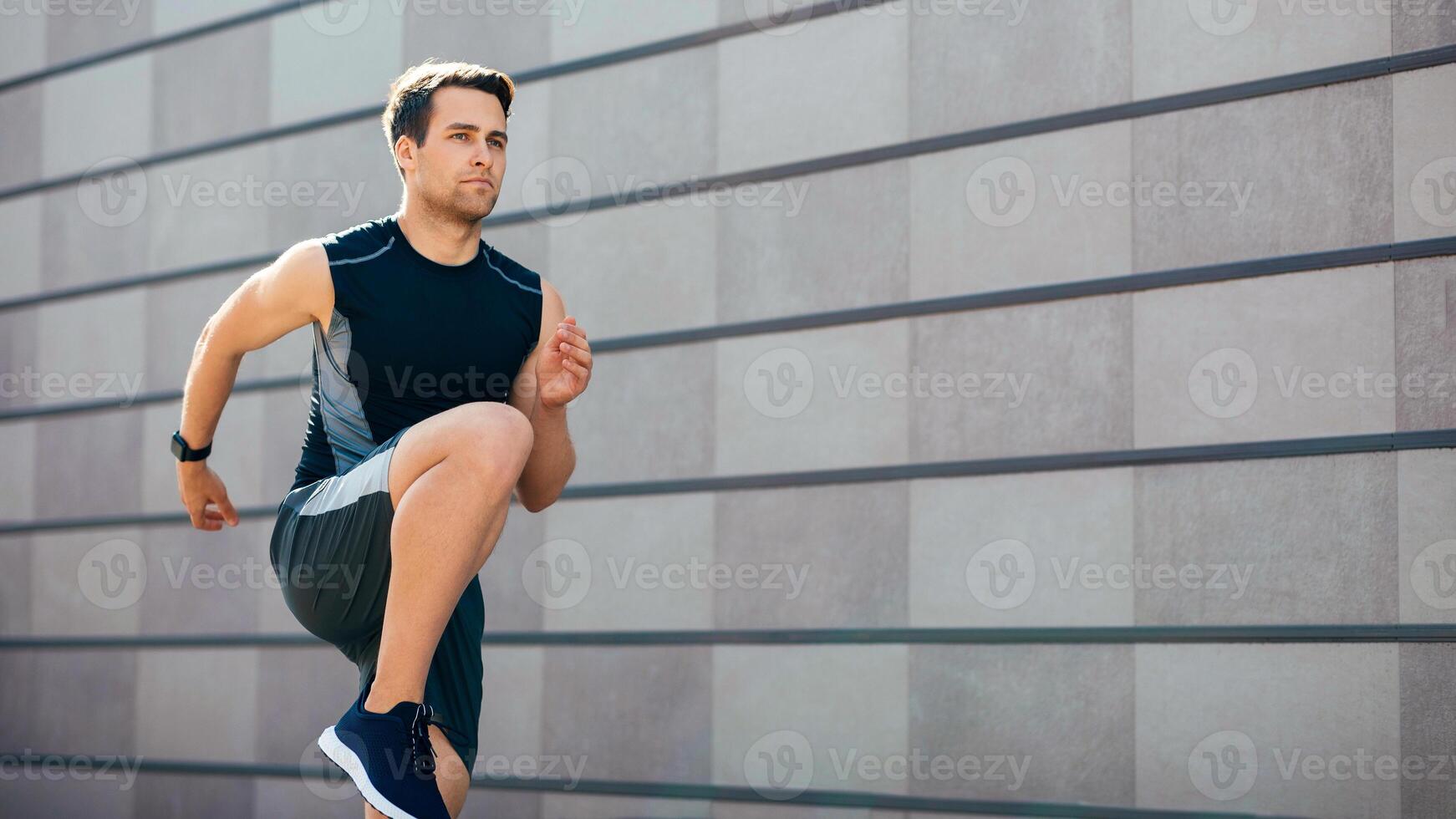 Serious athlete played sport in city. Young man raises legs high, running near gray wall, free space photo