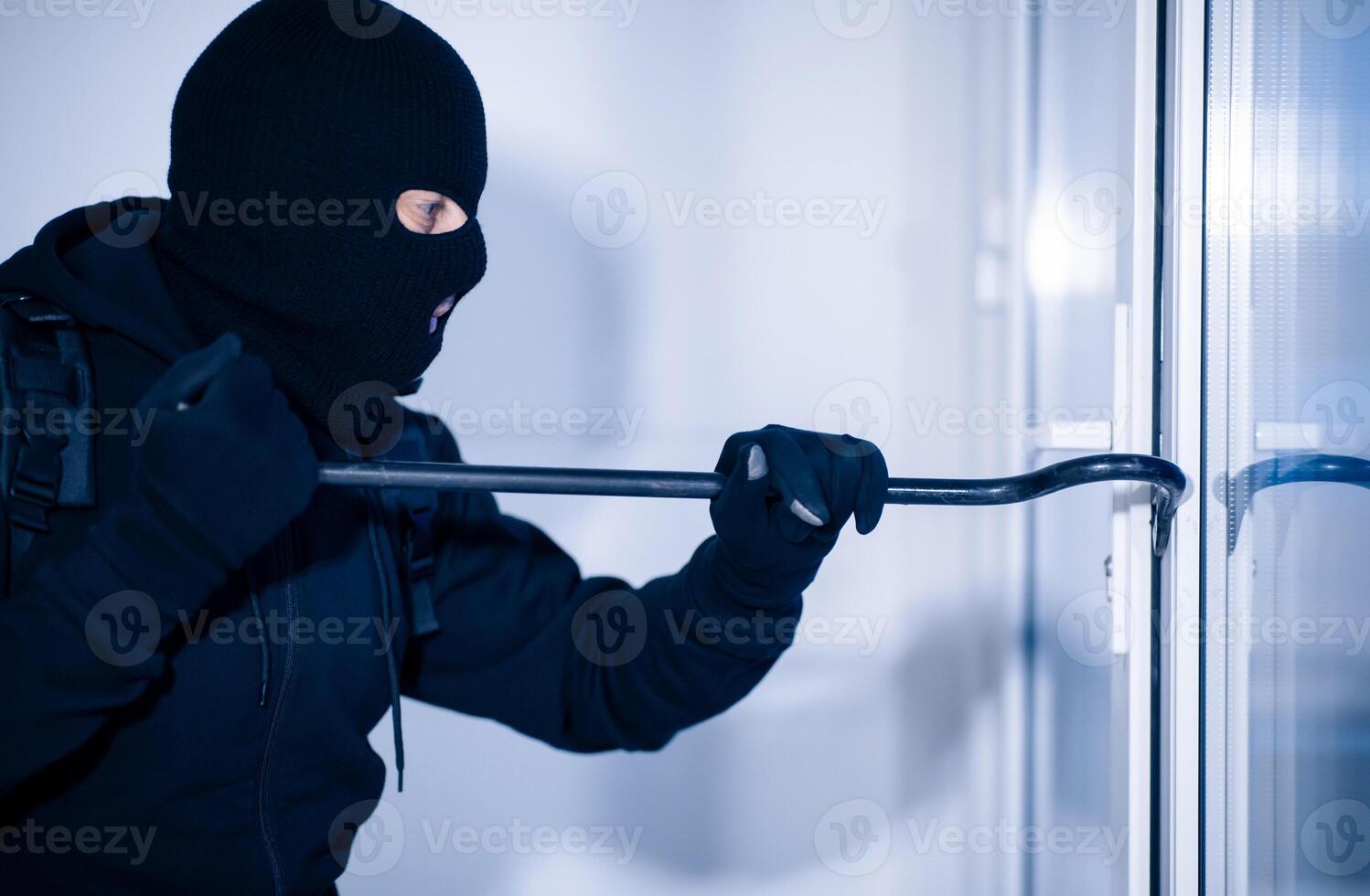 Intrusion Concept. Side view portrait of disguised prowler breaking house or office door with crowbar photo