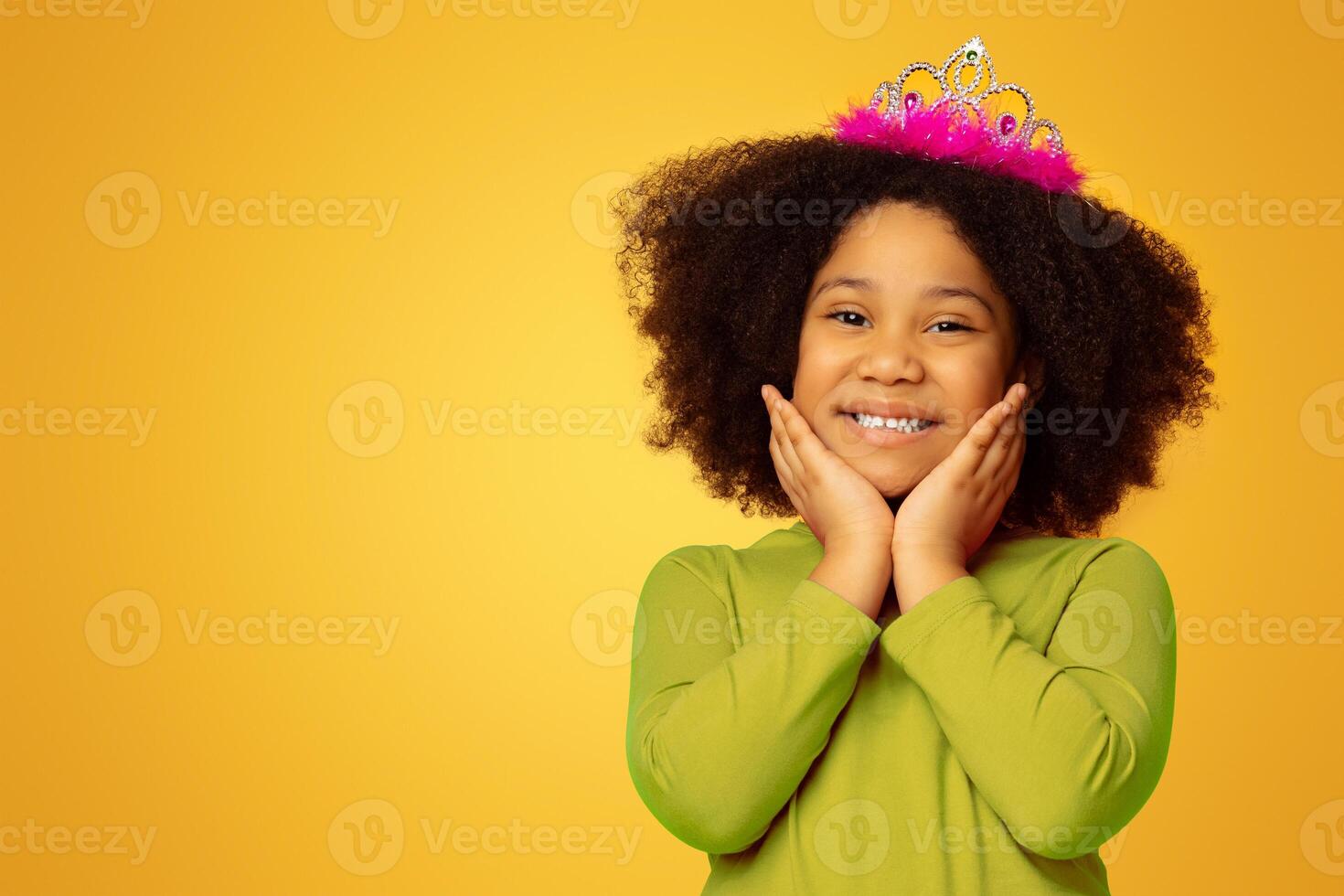 Little Princess. Portrait Of Cute Black Girl Posing To Camera With Crown On Head And Touching Her Face, Yellow Studio Background With Free Space photo