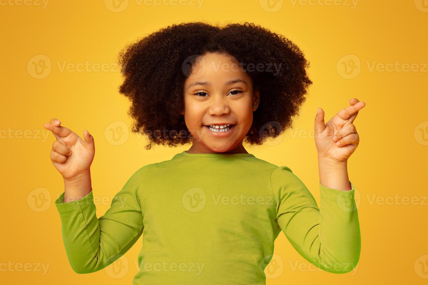 Making A Wish. Cute Little Afro Girl Pleading For Something With Crossed Fingers, Posing Over Yellow Background, Free Space photo