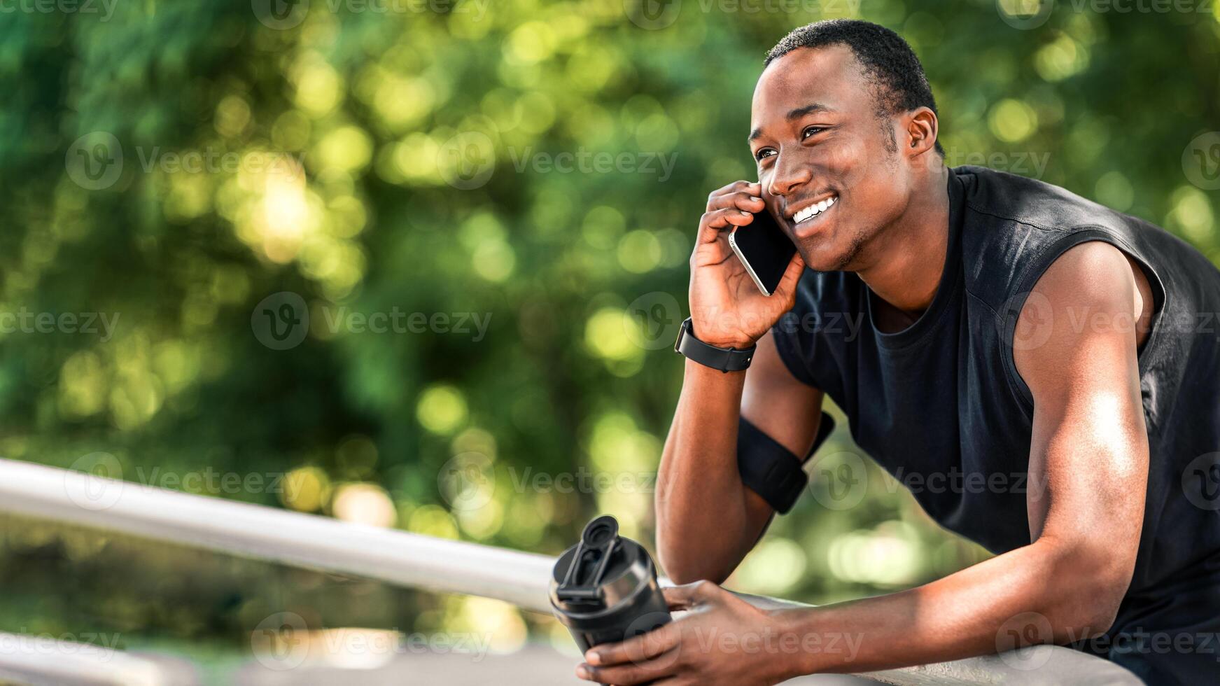 Excited african guy talking on phone during break, holding bottle with protein drink, exercising at park, free space photo