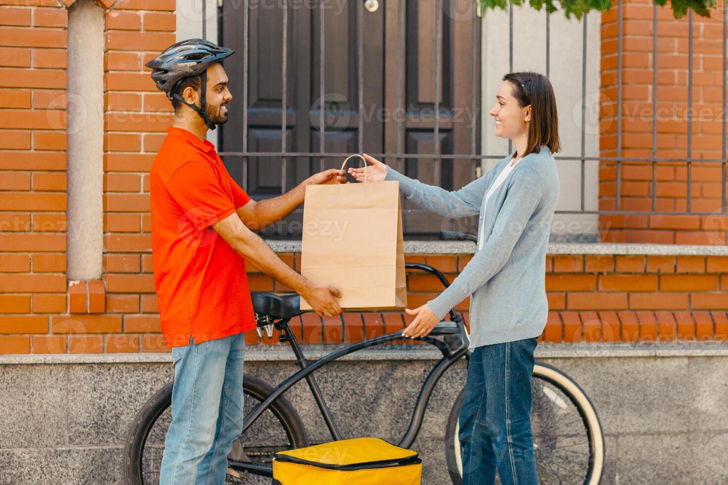 Safe delivery of purchase. Smiling courier in helmet, with big yellow backpack with bicycle, gives paper bag for client at front door, outdoors, side view, free space photo