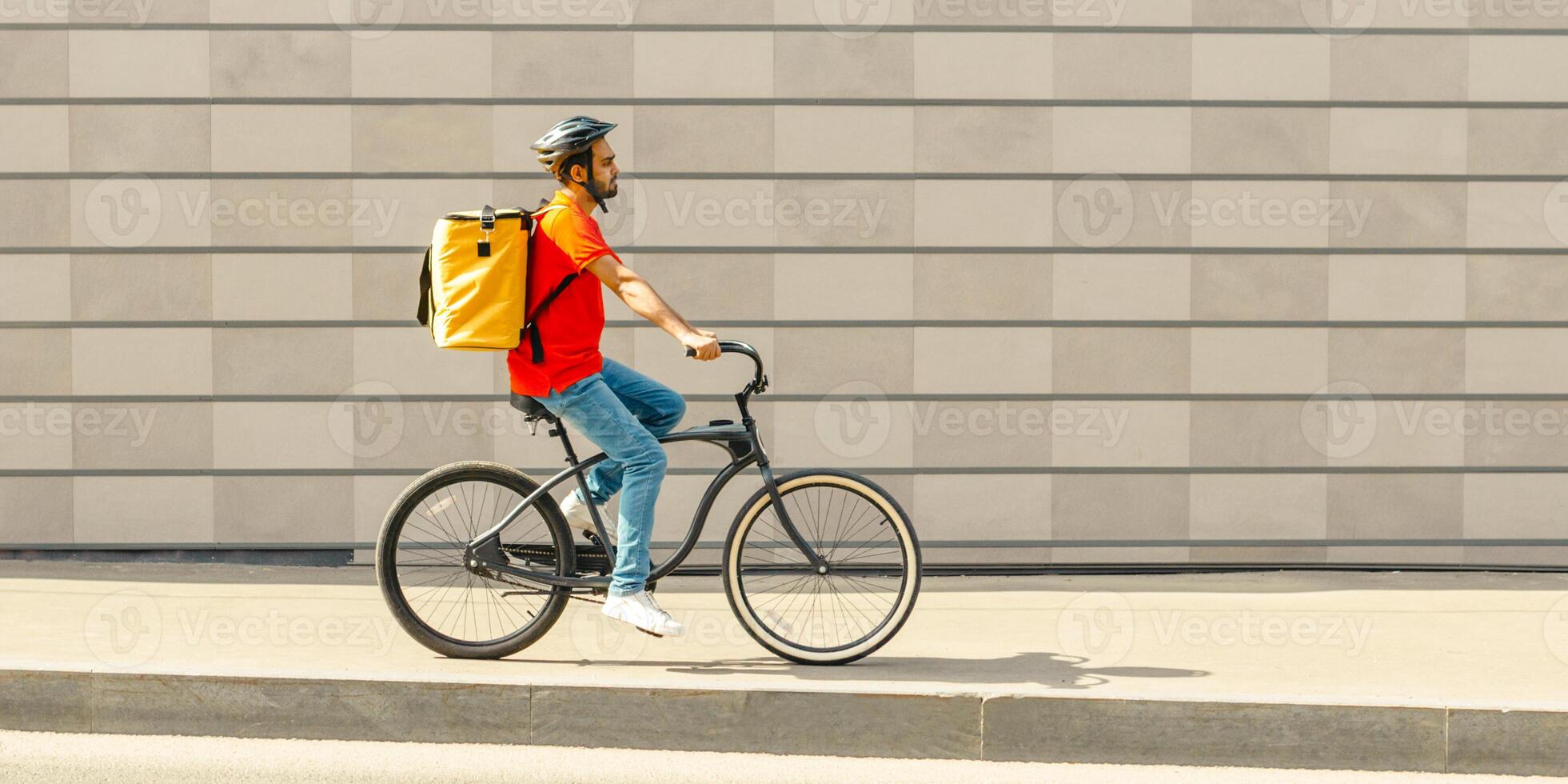 Deliveryman on bicycle. Young guy with beard and big backpack rides along path in city on gray brick wall background, side view, free space photo