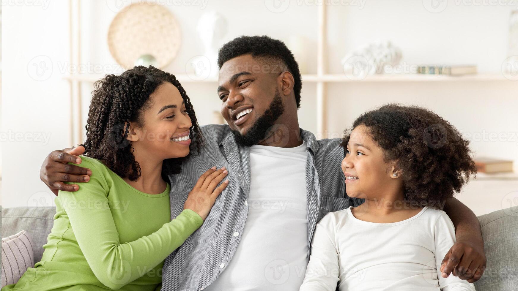 A joyful black family sits closely on a couch in their living room, sharing smiles and laughter. The warm afternoon light enhances their bond and creates a delightful atmosphere. photo