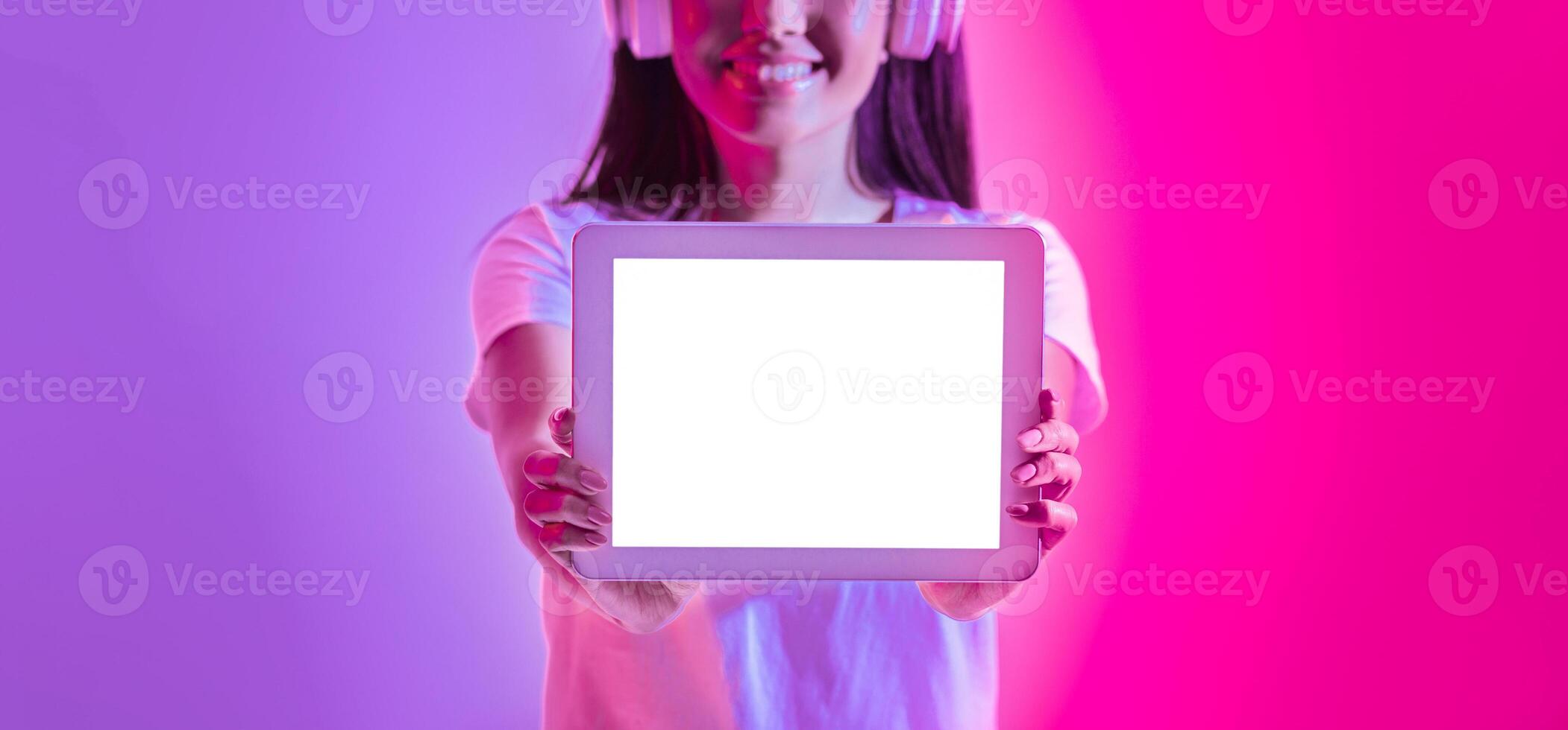 A smiling young woman wearing headphones holds a blank tablet in front of her against a vibrant pink and purple background. The atmosphere is lively and energetic. photo