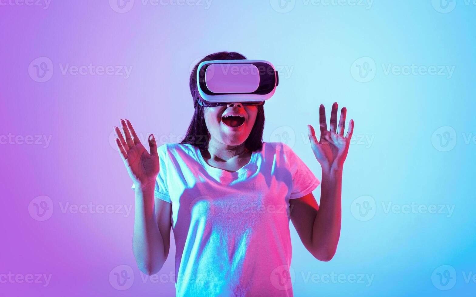 A young woman immersed in virtual reality shows excitement with her hands raised. The vibrant blue and purple lighting creates a captivating atmosphere, highlighting her joyful expression. photo