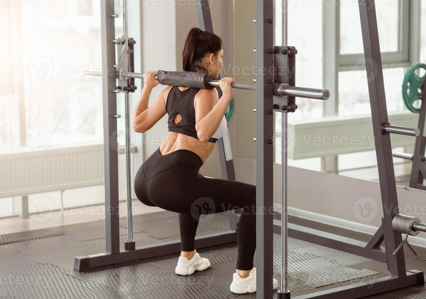 A woman in athletic wear demonstrates proper squat technique using a barbell in a gym setting. The well-lit space emphasizes a focus on strength training in the morning. photo