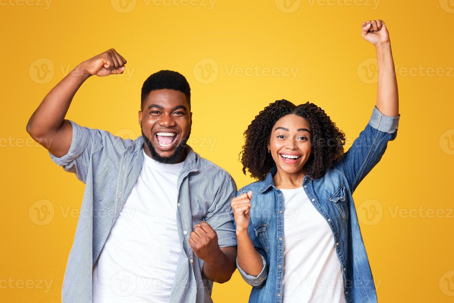 A man and woman joyfully celebrate, raising their fists in excitement. They have broad smiles and are dressed casually, standing against a vibrant yellow background. photo