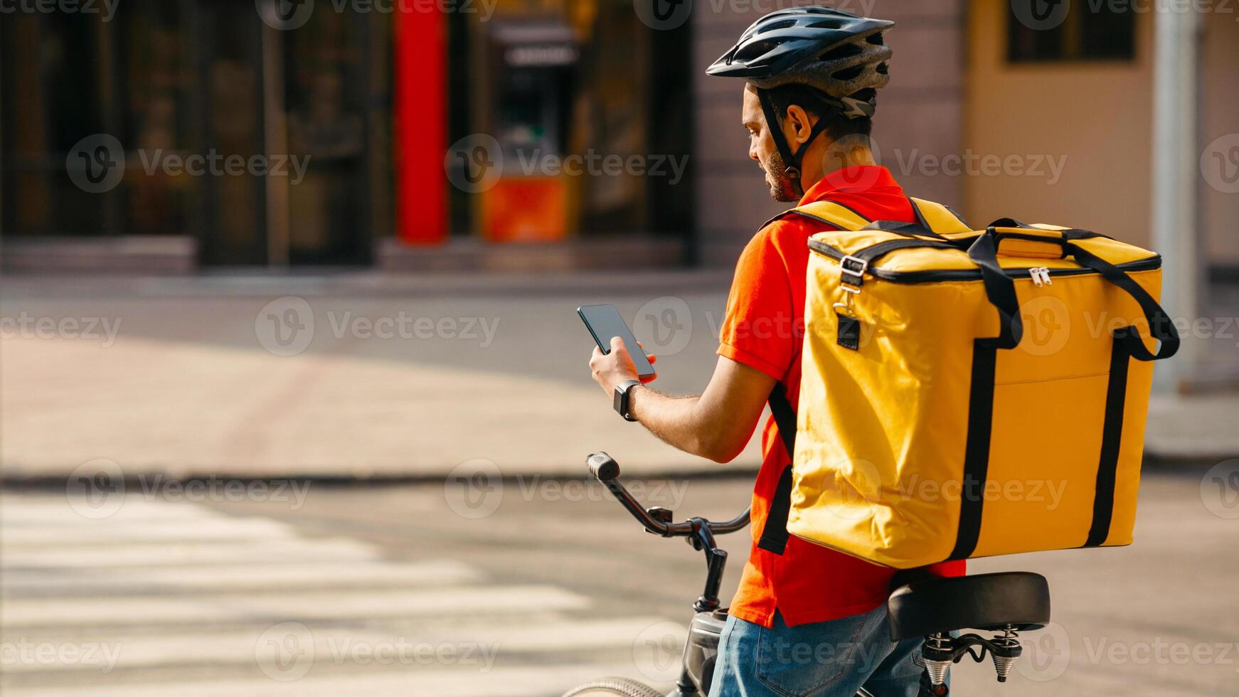 Young delivery man on bicycle. Guy with yellow backpack and helmet stopped at crossing and looked at phone, in city, free space photo