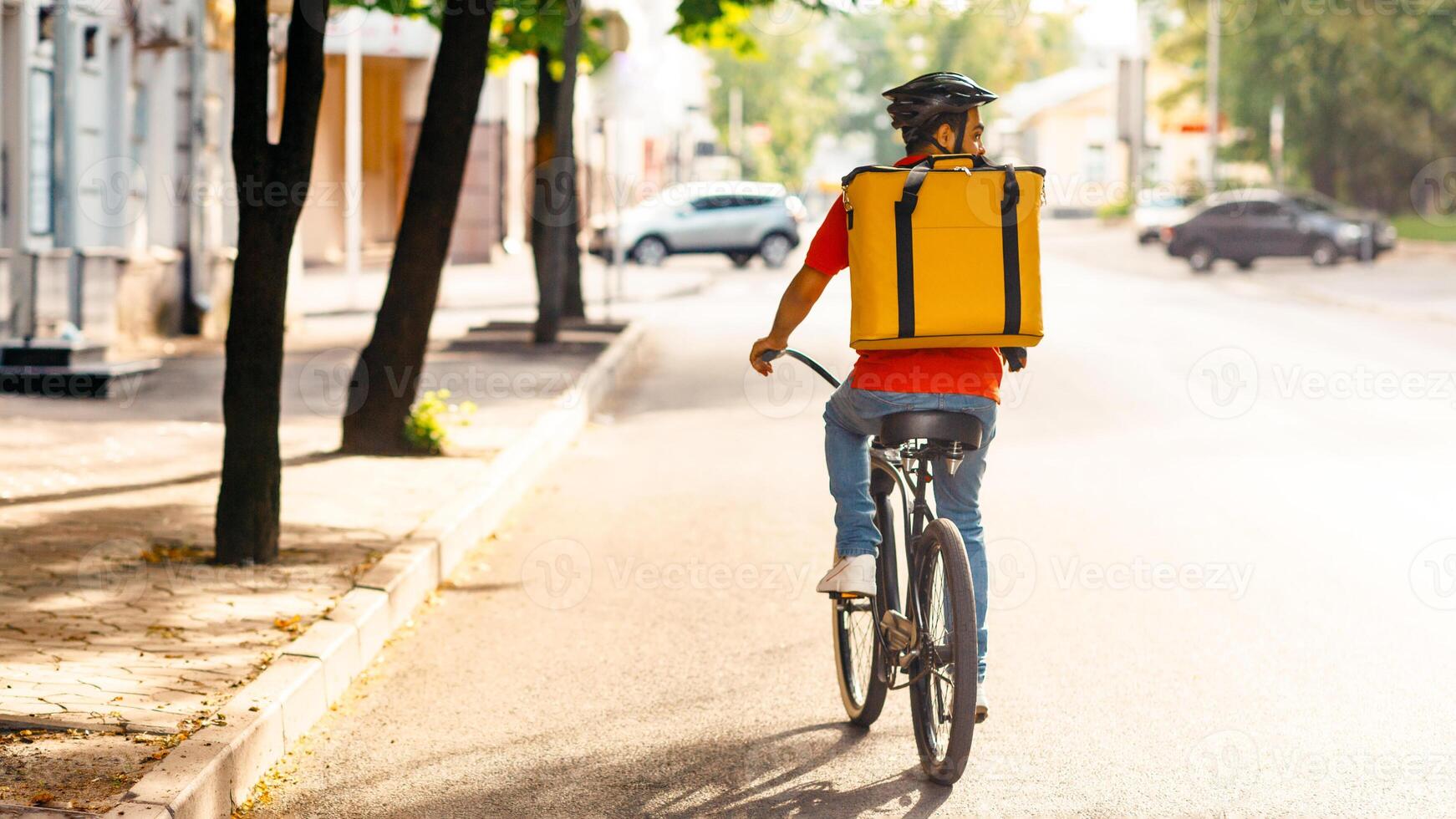 Courier on bicycle with parcel, bike, delivery service. Young guy in uniform, protective helmet with yellow thermo bag riders on road in city in summer and searches client address, empty space, back photo