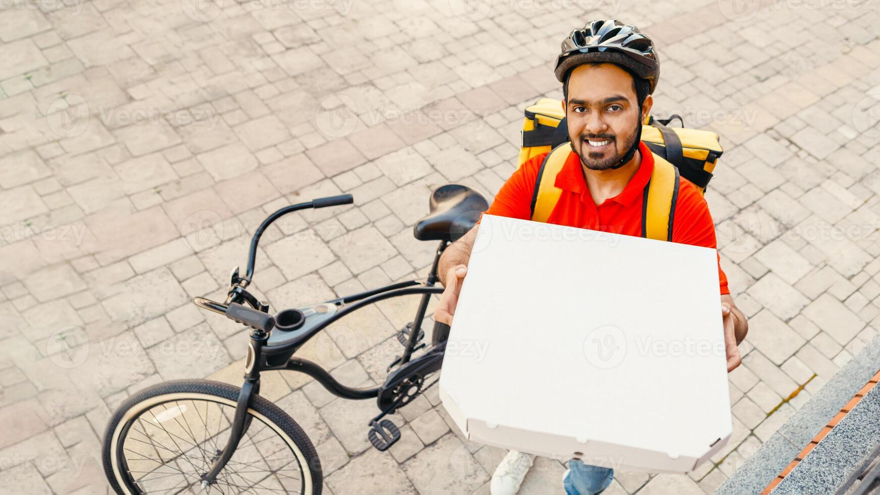 Order delivery. Smiling young man with beard, in helmet, shows box of pizza to peephole, near bike on street, free space photo