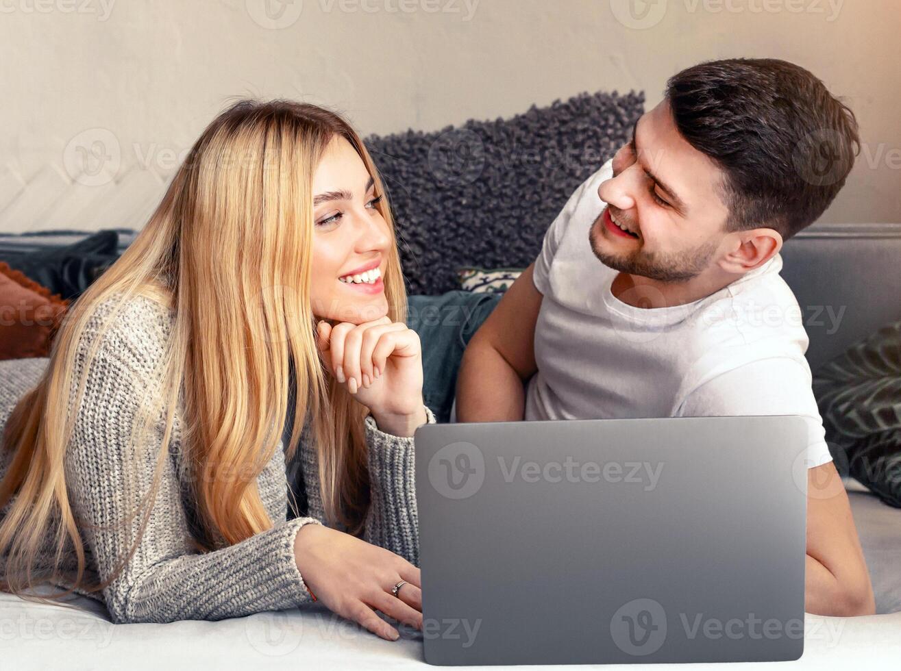 A young couple lies comfortably on a couch, smiling and looking at a laptop together. They share a cozy moment, engaging with each other and the content on the screen. photo