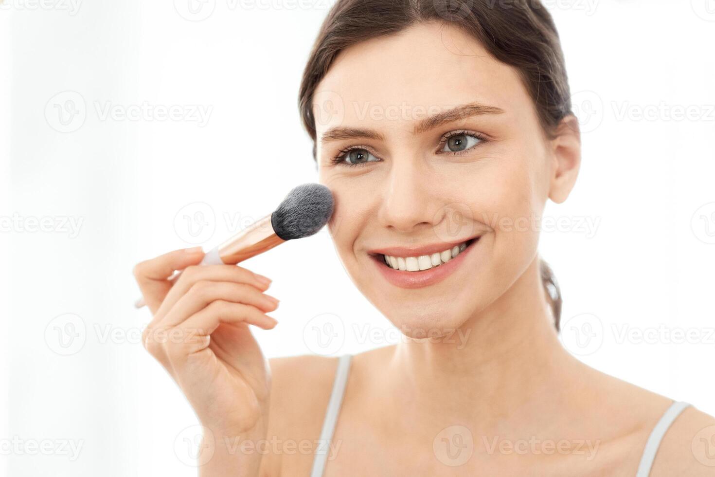Closeup of attractive young woman applying blush on her face, sitting in front of mirror, copy space. Smiling attractive young lady doing makeup before going out, shot over shoulder in bedroom photo