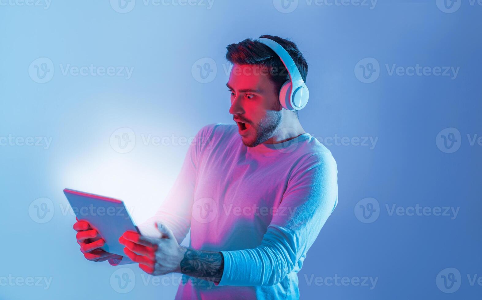 A young man wearing headphones shows a surprised expression while holding a tablet. The background features captivating blue and pink lighting, enhancing the vibrant atmosphere. photo