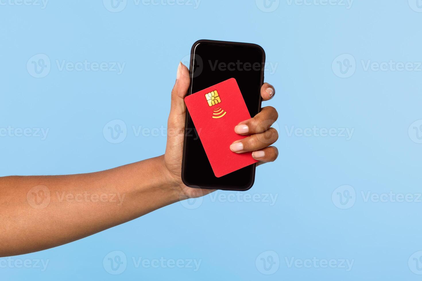 An African American woman confidently displays her mobile payment method using a smartphone and a contactless card. The vibrant blue background highlights her modern lifestyle. photo