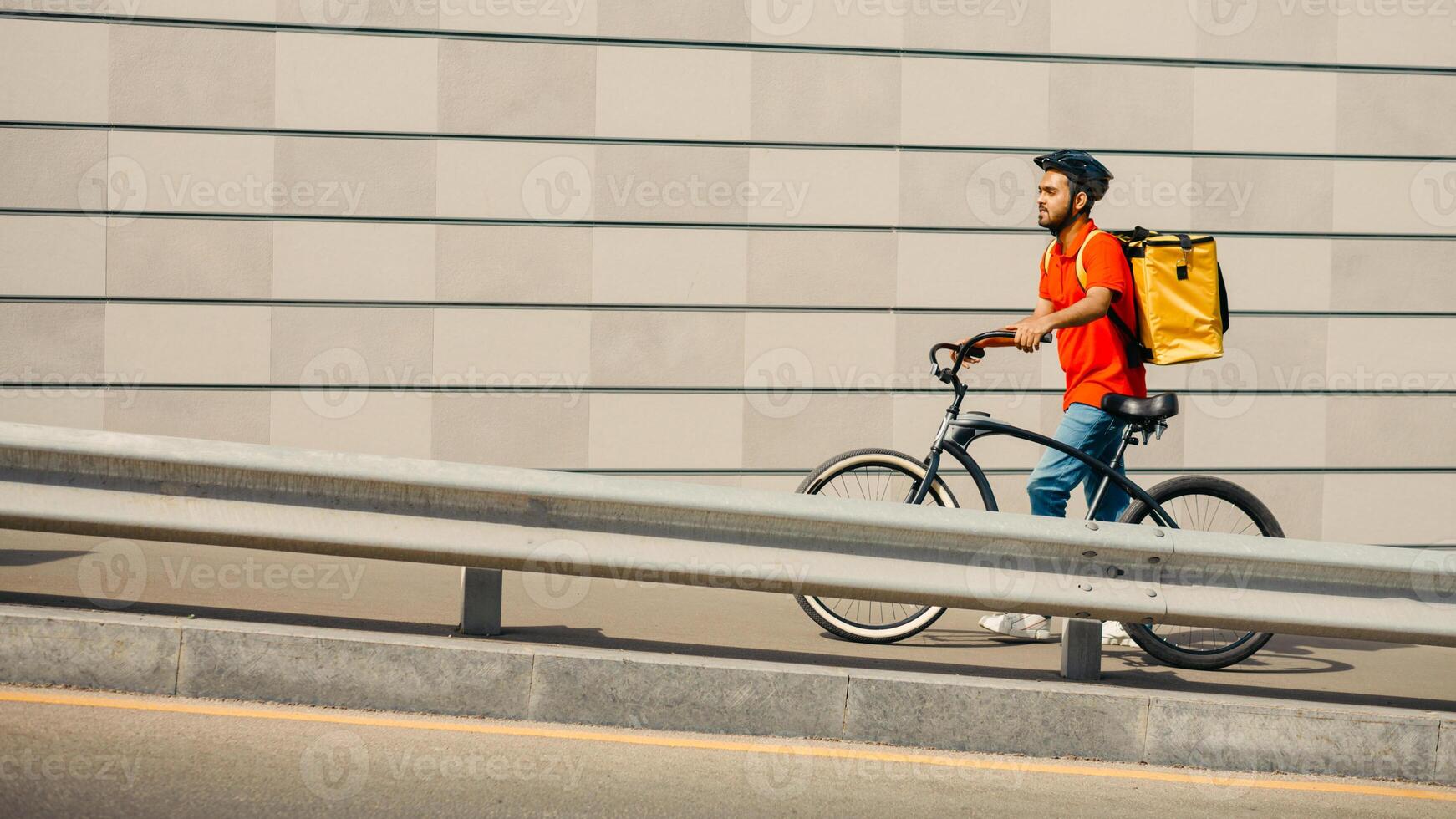 Road to client with online order. Courier in protective helmet with yellow backpack and bicycle going up the street on gray wall background, side view, free space photo