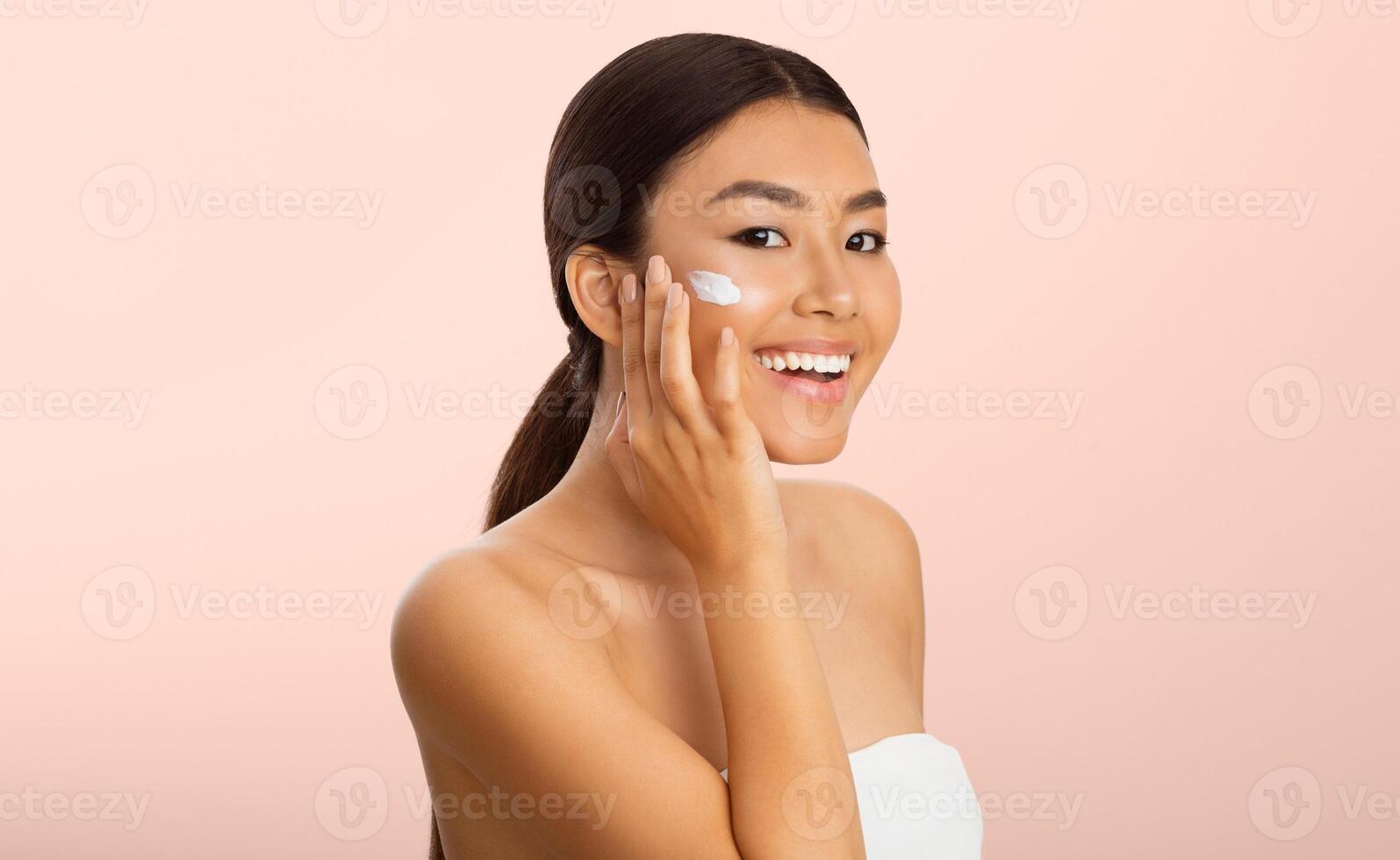 Beauty Concept. Asian Woman Applying Moisturizer On Face, Pink Studio Background photo