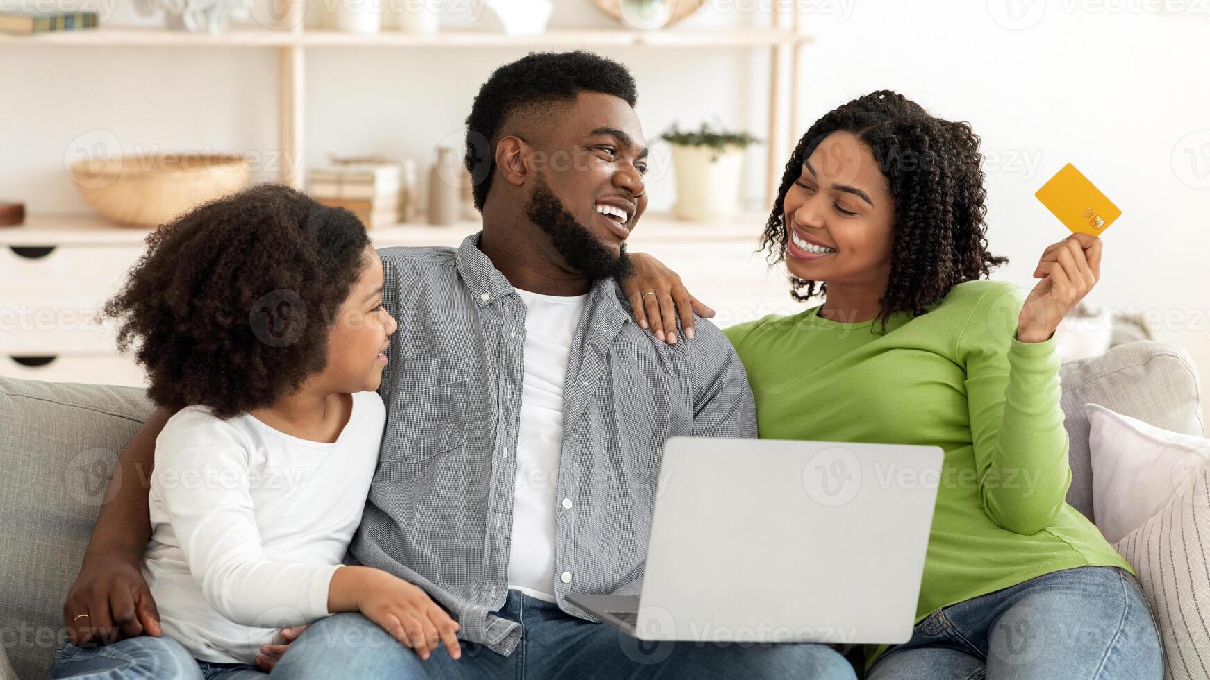 A black family is gathered in their cozy living room, happily engaging in online shopping while sharing smiles and laughter. The child sits beside the parents, creating a warm atmosphere. photo