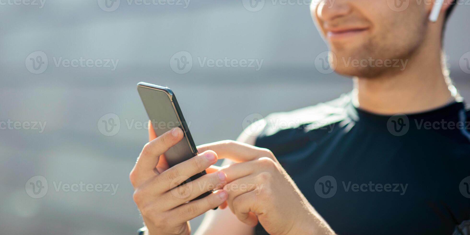 Favorite track for training and great mood. Smiling guy in sportswear with wireless headphones looks at smartphone, close up, free space, sun flare photo