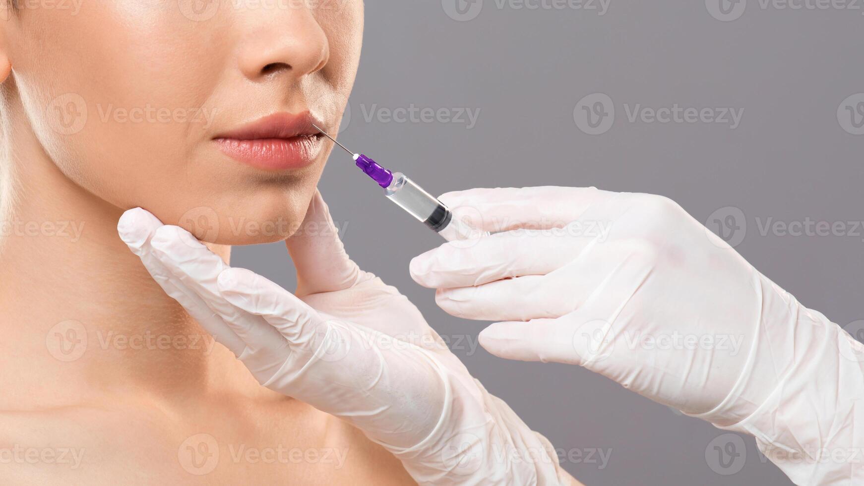 Aesthetic modern medicine concept. Lip injection for young woman over grey studio background, closeup. Unrecognizable cosmetitian injecting lip filler for beautiful lady looking at copy space photo