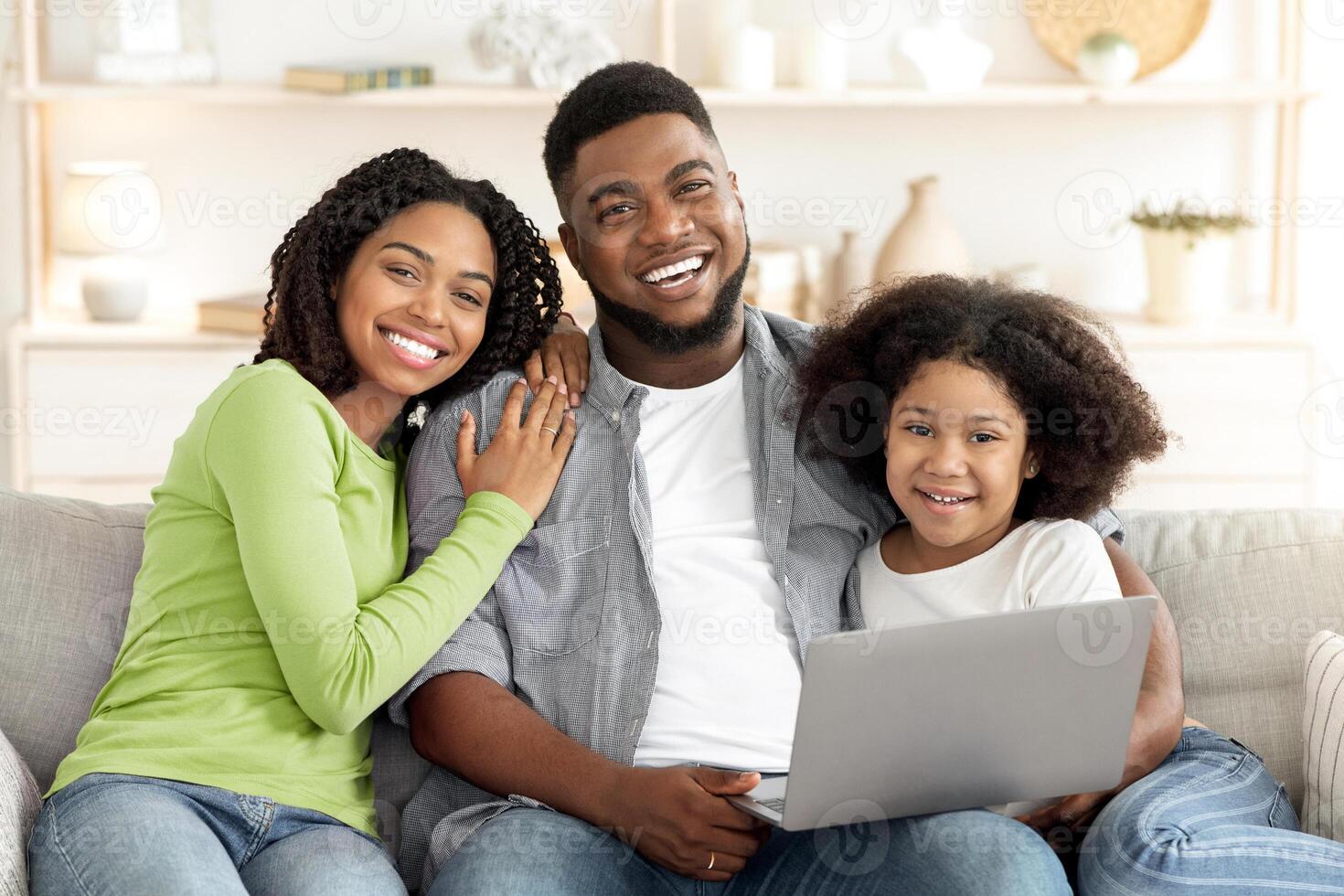 A smiling black family of three gathers on a couch in their cozy living room, engaging with a laptop. They appear joyful, reflecting a warm and loving atmosphere during family time. photo