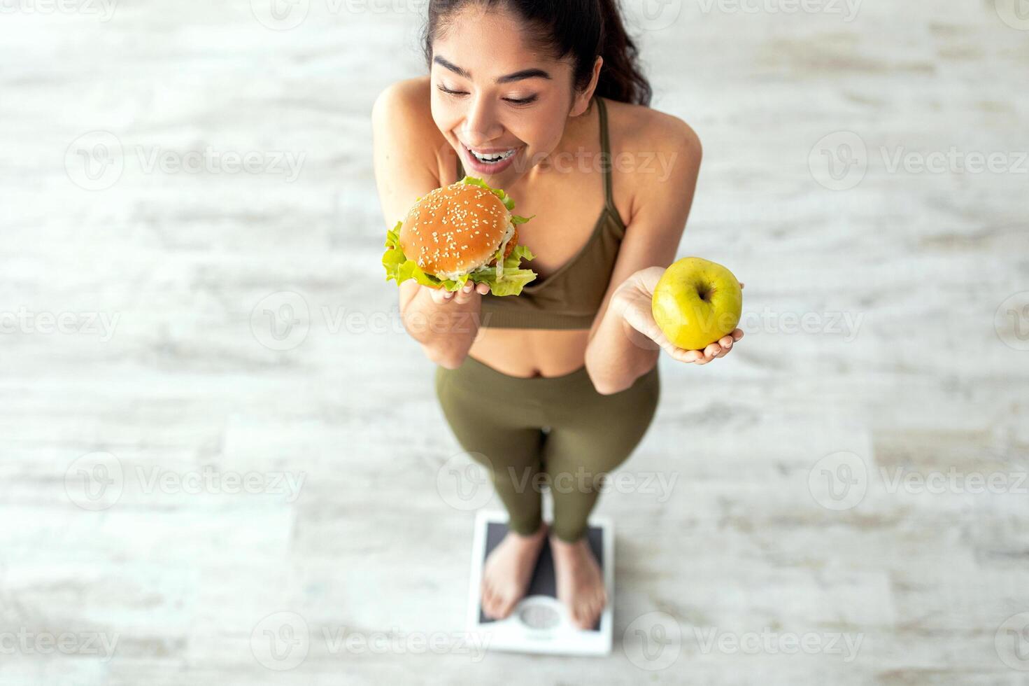 A young Indian woman smiling while holding a burger in one hand and an apple in the other. She stands on a scale, representing the balance between fitness and healthy eating at home. photo