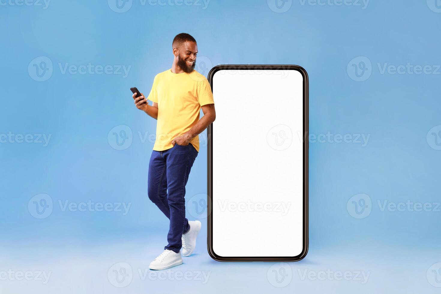 A millennial African American man stands casually with a smartphone in hand, smiling at the screen. He is dressed in a bright yellow shirt and blue pants, with a large blank phone beside him. photo
