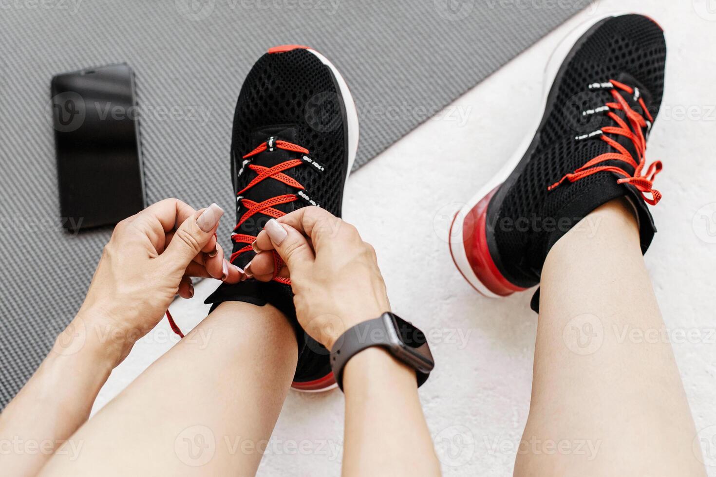Athletic female tying laces on shoes, taking break between training at home. On floor on mat bottle of water, headphones and smartphone with blank screen, top view, free space, cropped, studio shot photo