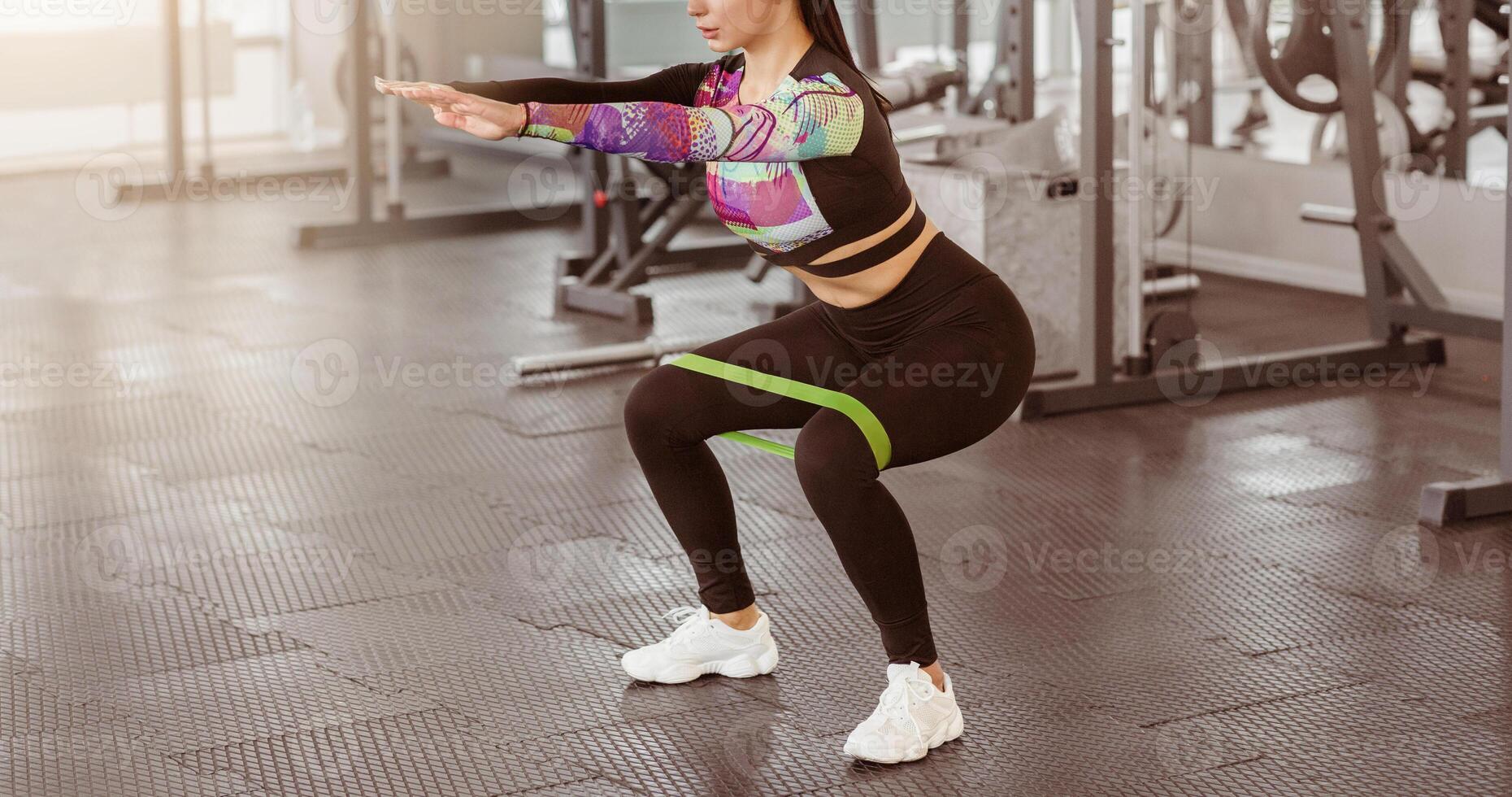 A woman is engaged in a squat exercise using a resistance band in a well-lit gym. She focuses on maintaining proper form while working out during a morning fitness session. photo