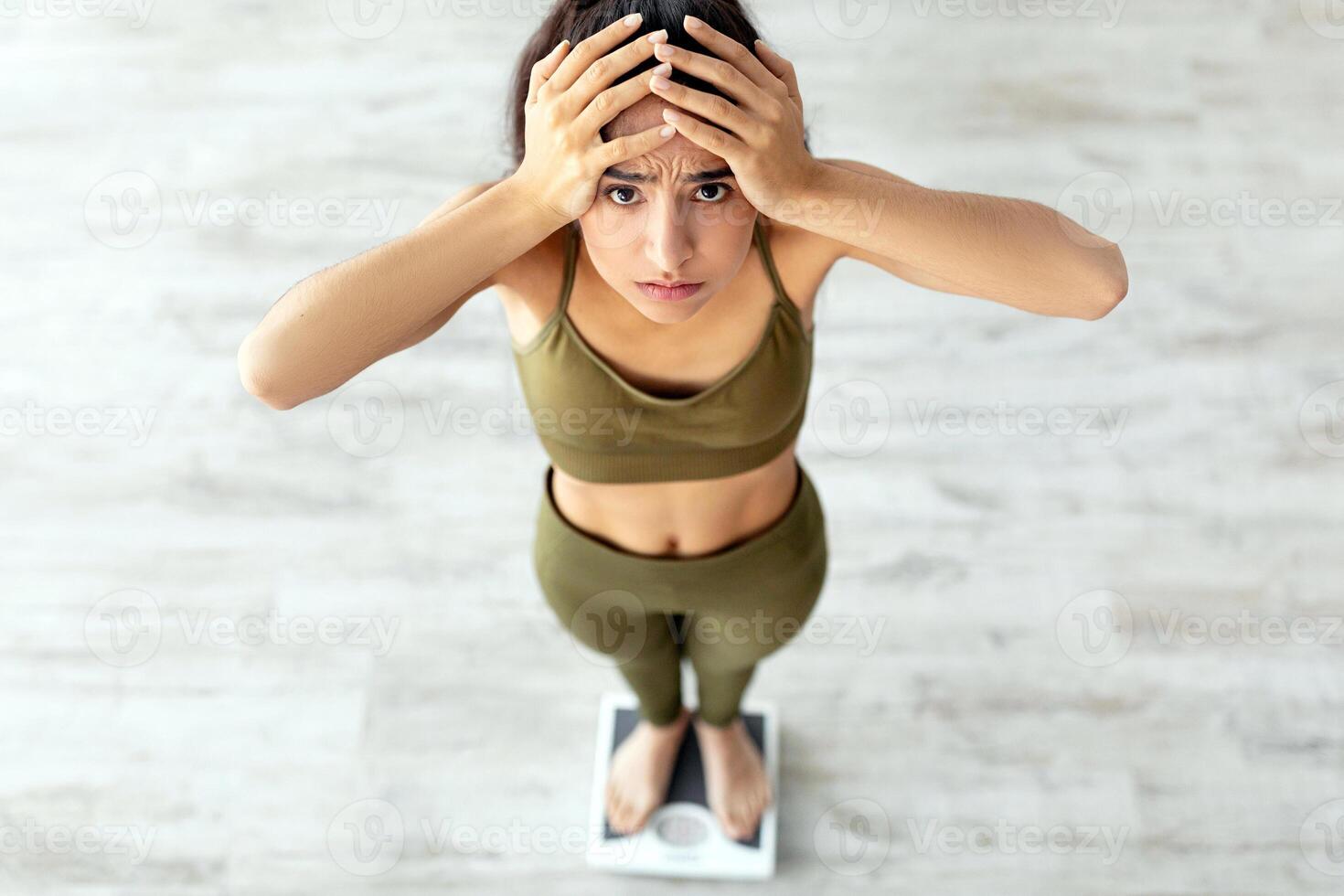 A young Indian woman stands on a scale at home, looking concerned and frustrated about her weight. She is wearing workout attire, reflecting a commitment to her fitness goals. photo