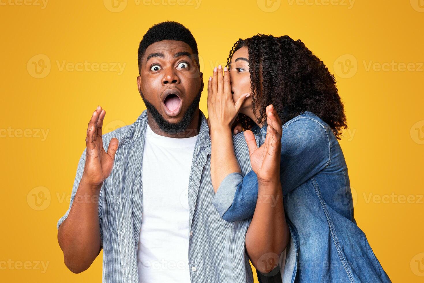 A woman leans in to whisper a secret to a shocked man, who is expressing surprise. The vibrant yellow background adds energy to the moment, highlighting their emotions. photo