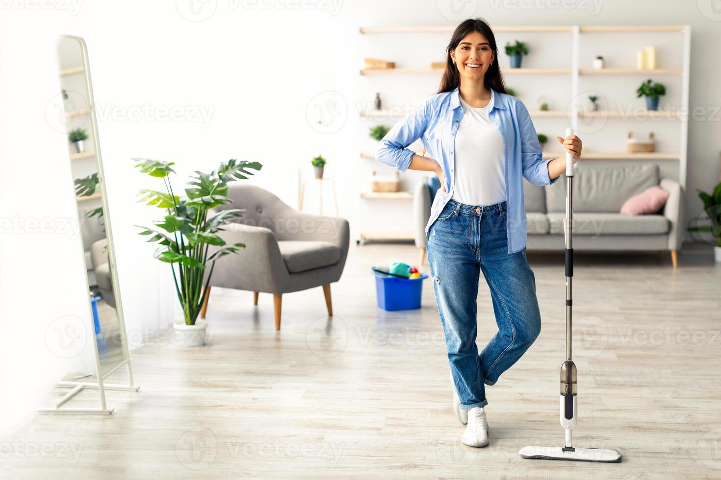 A young woman stands confidently in her stylish home, equipped with a mop. She smiles while tidying her living space, surrounded by modern furniture and greenery, copy space photo