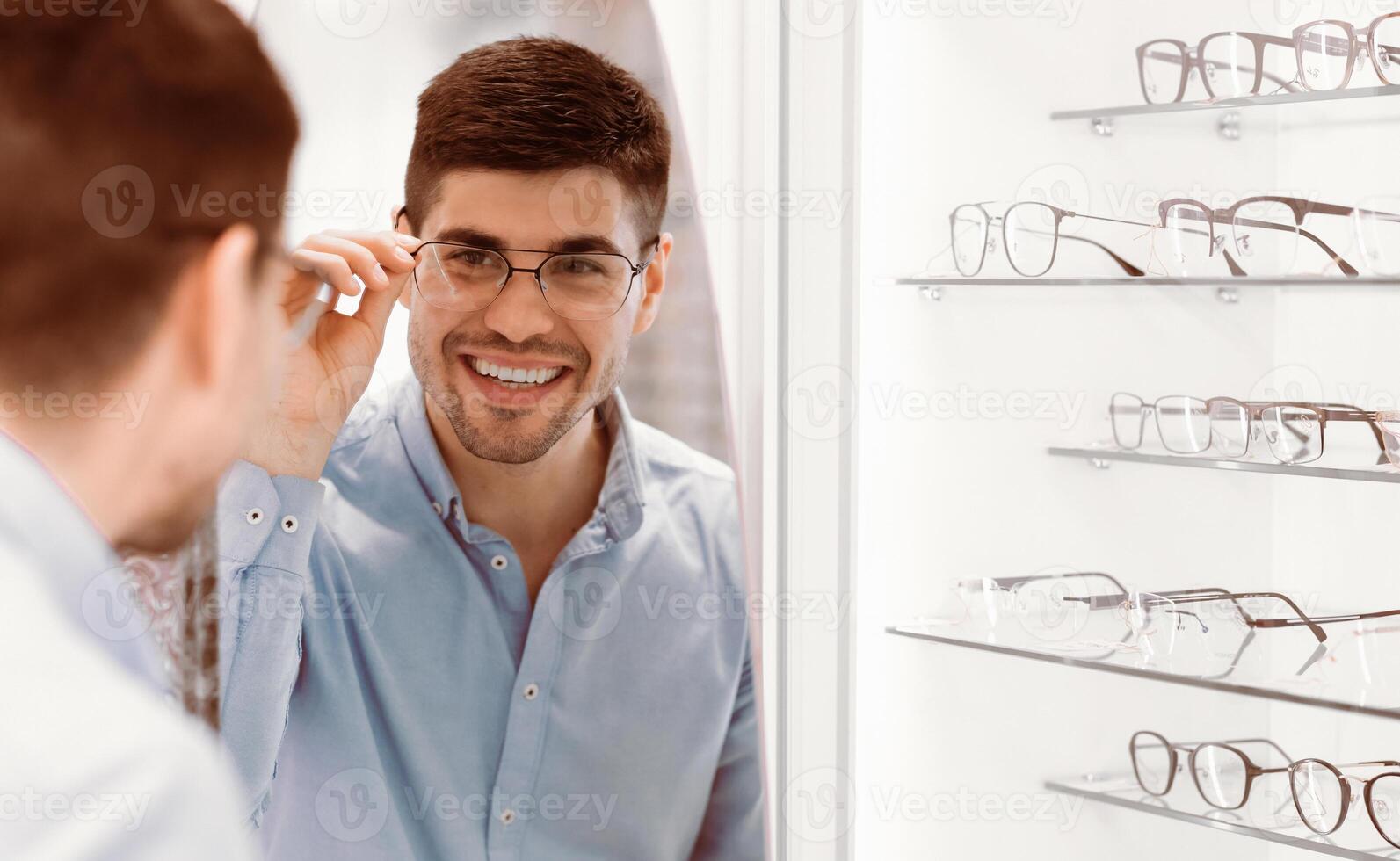 A young man examines himself in the mirror while trying on a stylish pair of glasses in an eyewear store, displaying a joyful expression as he checks the fit and style. photo