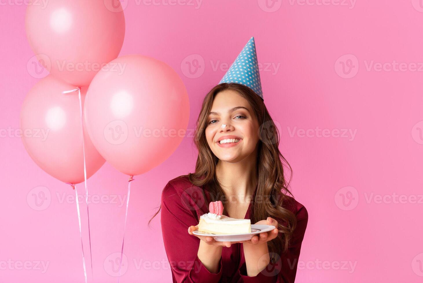 A joyful woman wearing a party hat holds a plate of cake, smiling against a bright pink background with balloons. The atmosphere radiates warmth and celebration during a birthday party. photo