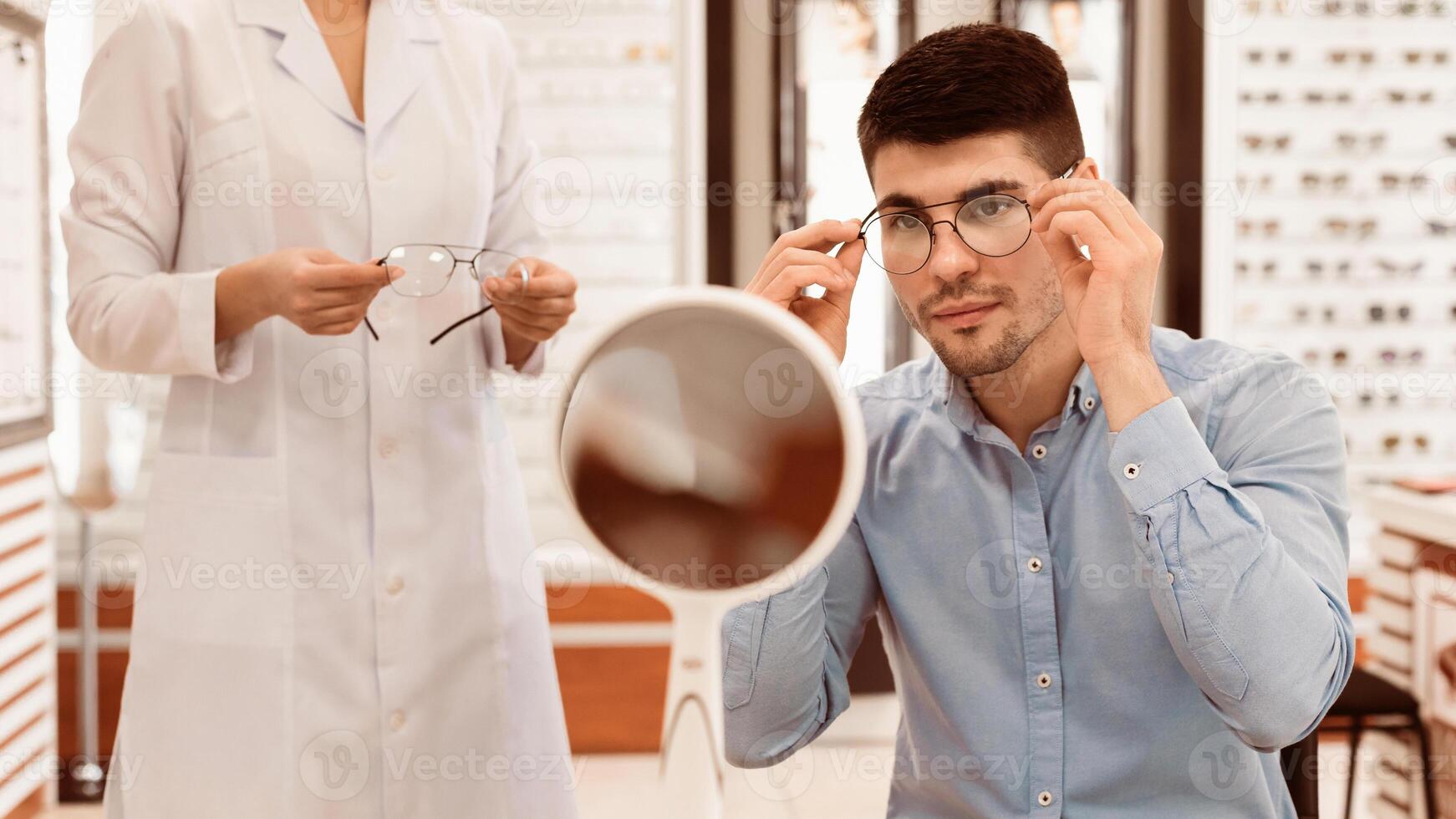 A young man adjusts the frame of glasses while looking in a mirror at an optical store. An optometrist holds a pair of glasses, assisting him in the selection process. photo