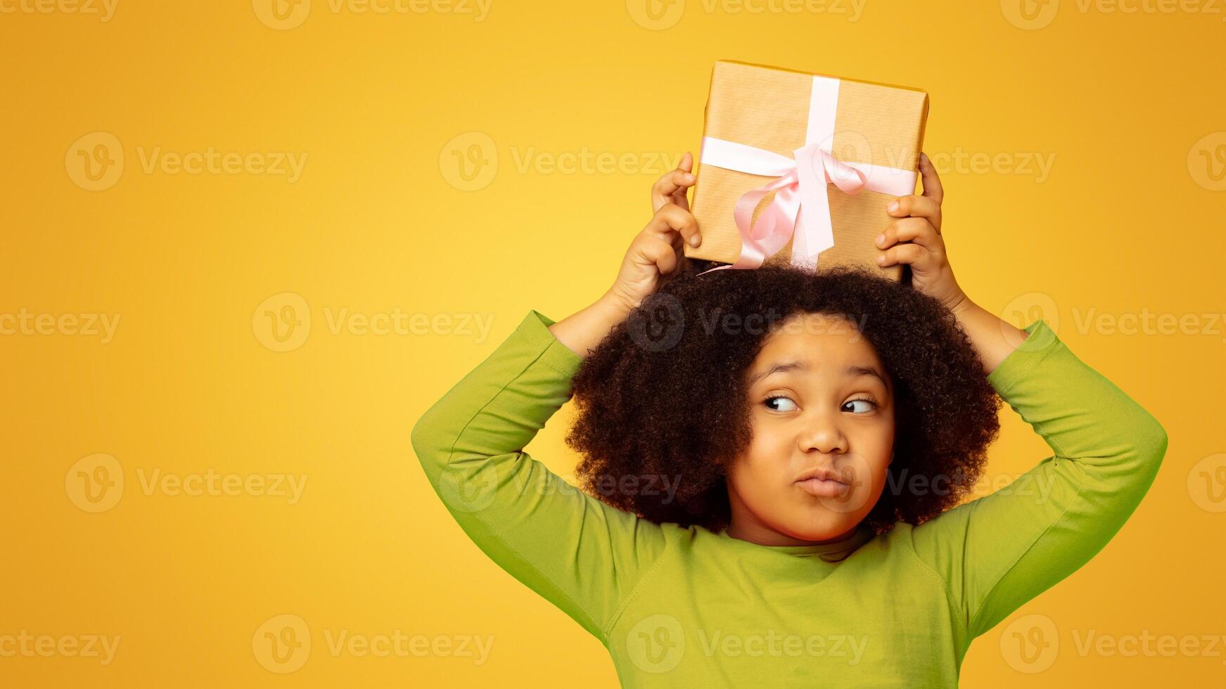 Funny little afro kid holding gift box above head, posing over yellow background with copy space, panorama photo