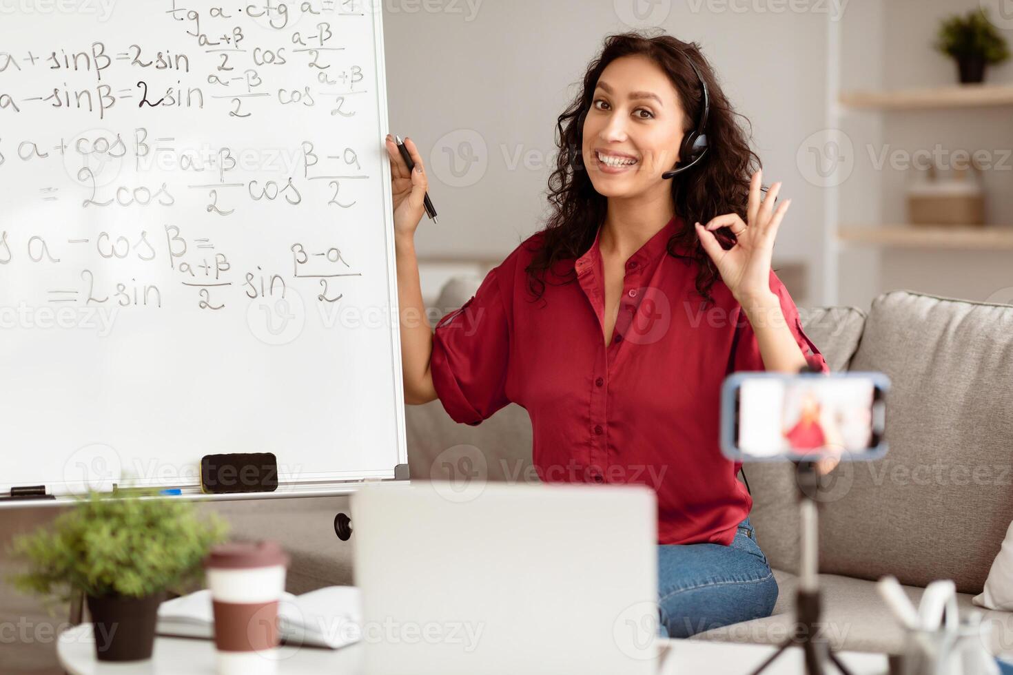 A young middle eastern woman is tutoring students remotely from her cozy living space. She stands next to a whiteboard filled with math equations, smiling and using a headset. photo