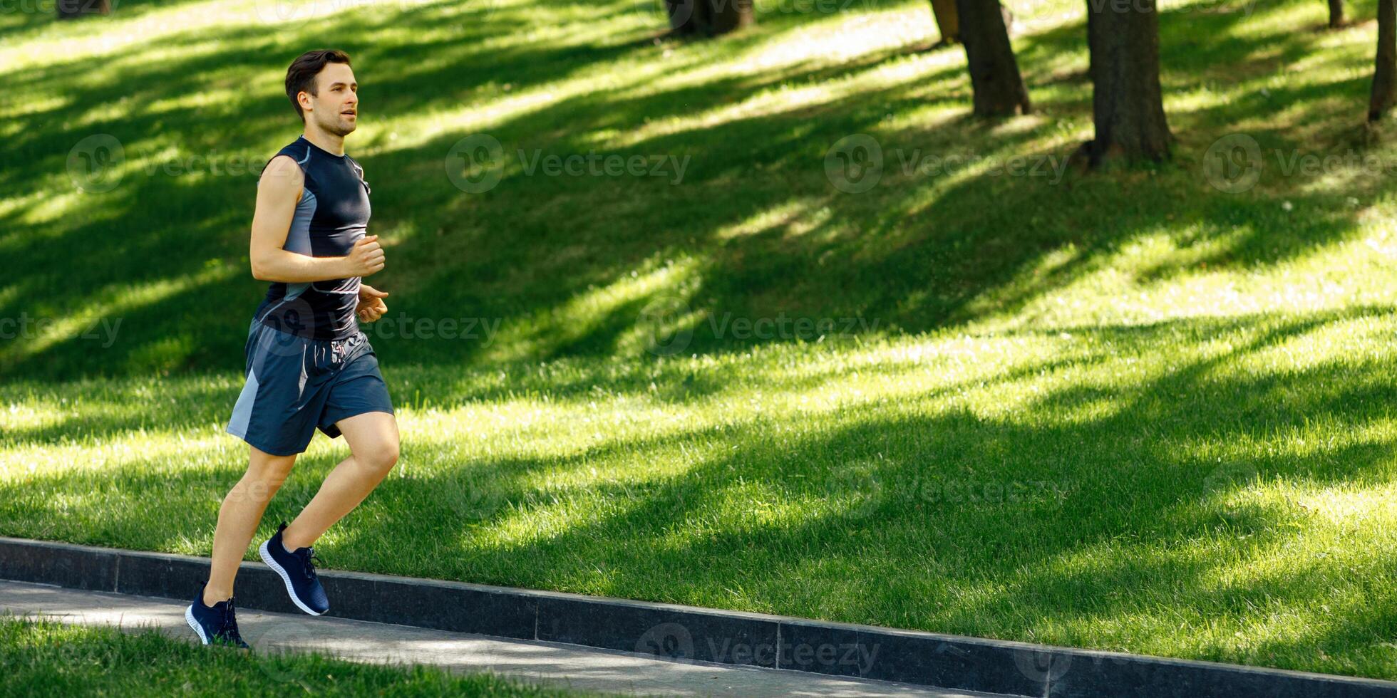 Physical activity outside. Guy in sportswear with sneakers and fitness tracker runs at path in park, free space, sun flare photo