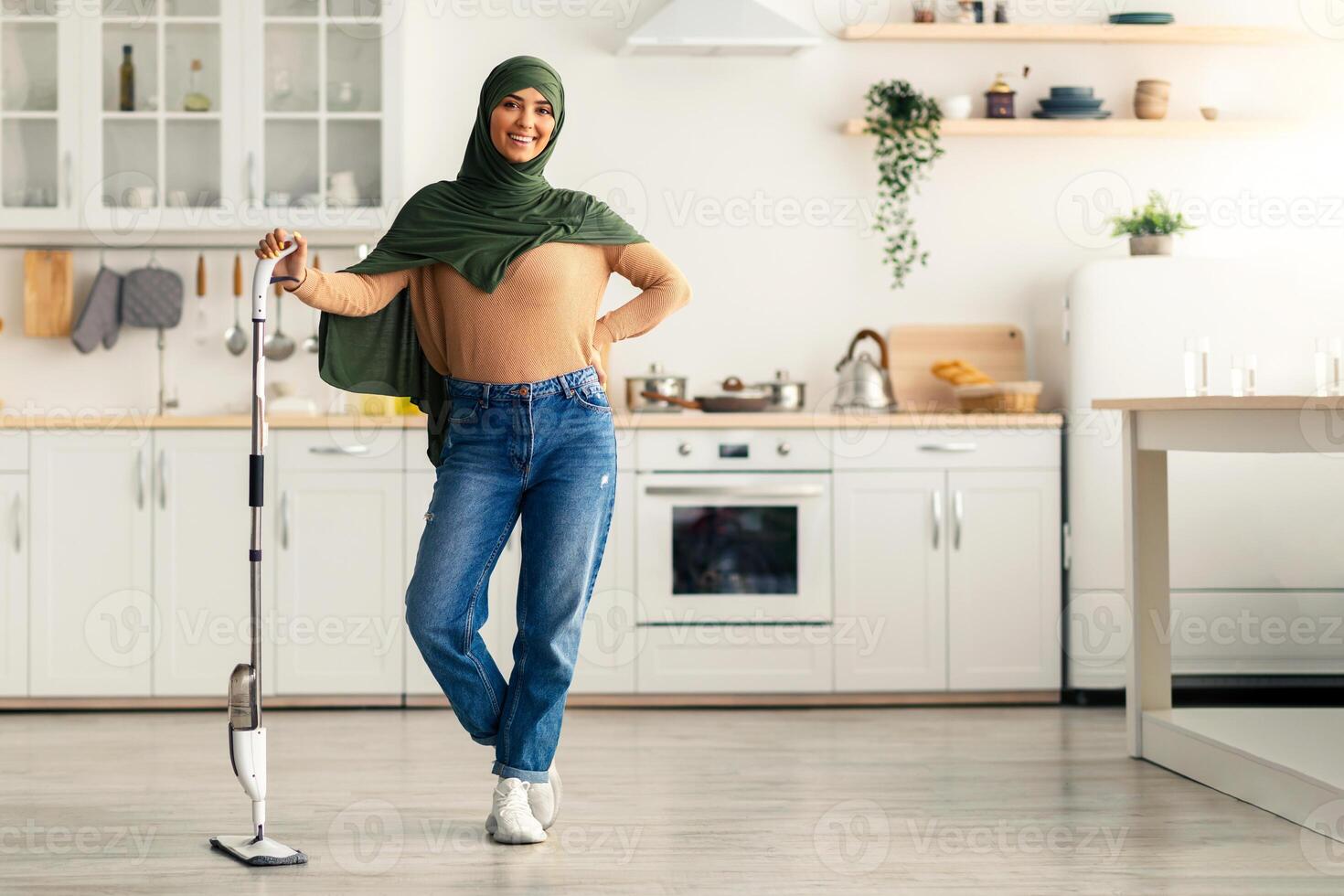A young woman stands in a bright, modern kitchen, smiling while holding a cleaning device. She is dressed casually and appears relaxed during her housekeeping task in a vibrant home. photo