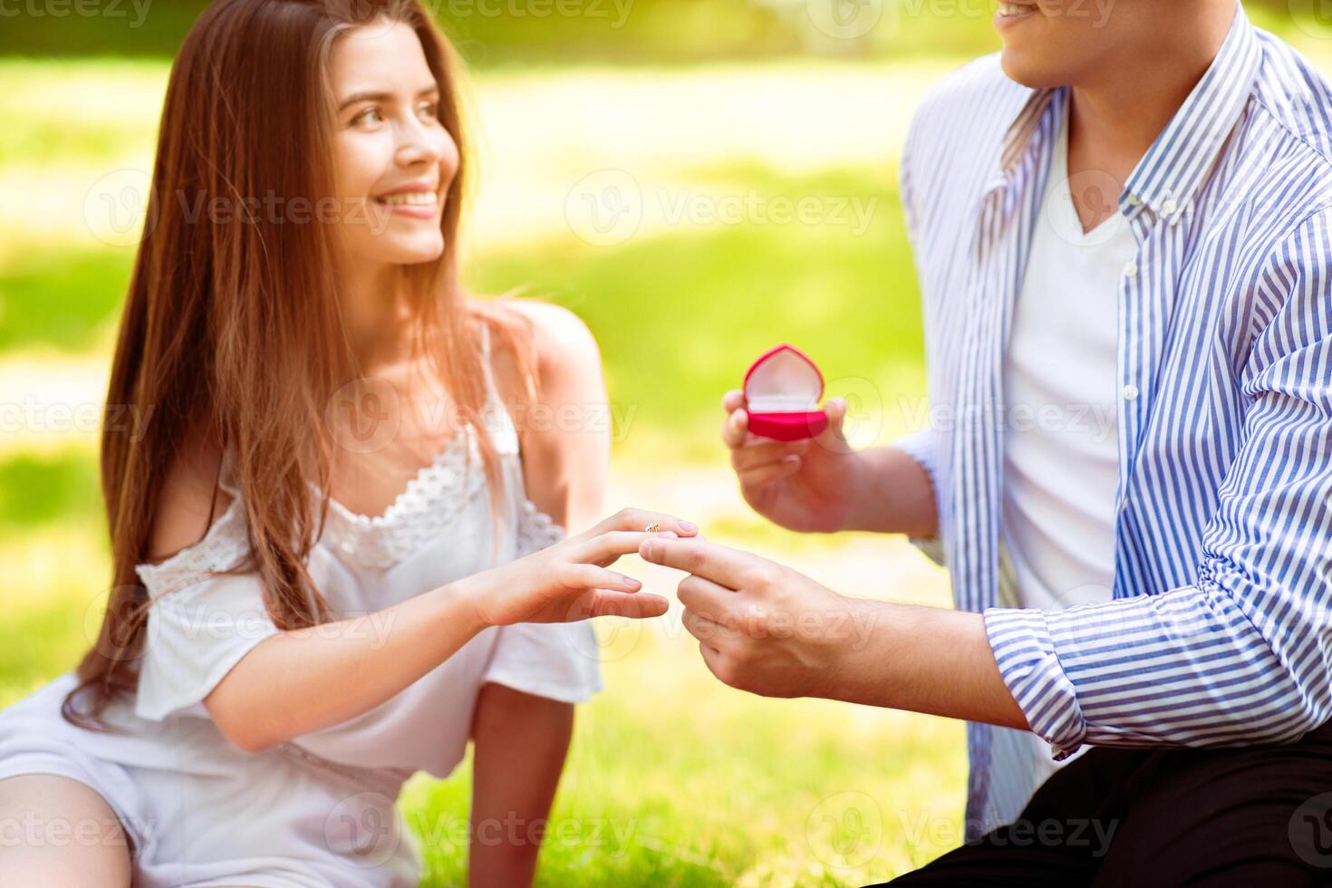 Romantic proposal. Millennial man putting ring on his beautiful girlfriend's finger at park photo