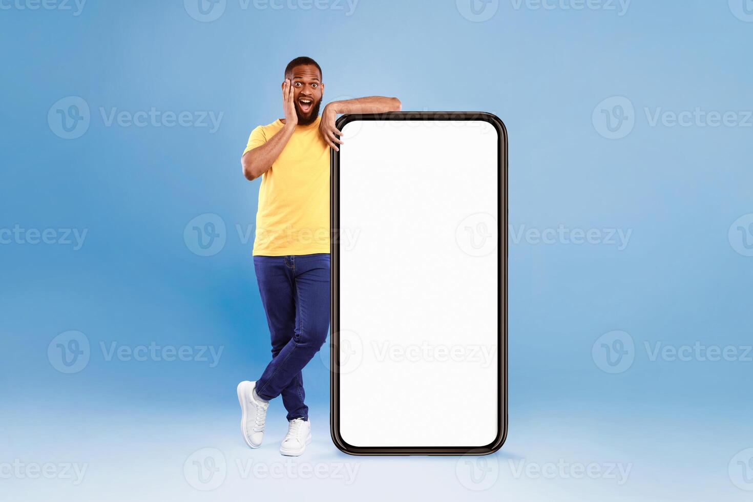 A joyful African American man poses next to an oversized blank smartphone. He appears enthusiastic and happy, showcasing a vibrant lifestyle in a bright studio. photo