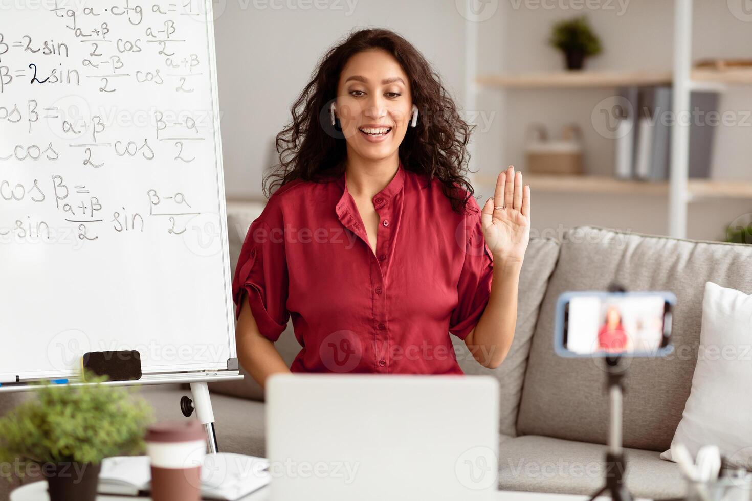 A millennial Arab woman smiles while teaching math remotely. She gestures as she engages with students on a call, surrounded by a friendly workspace with a whiteboard. photo