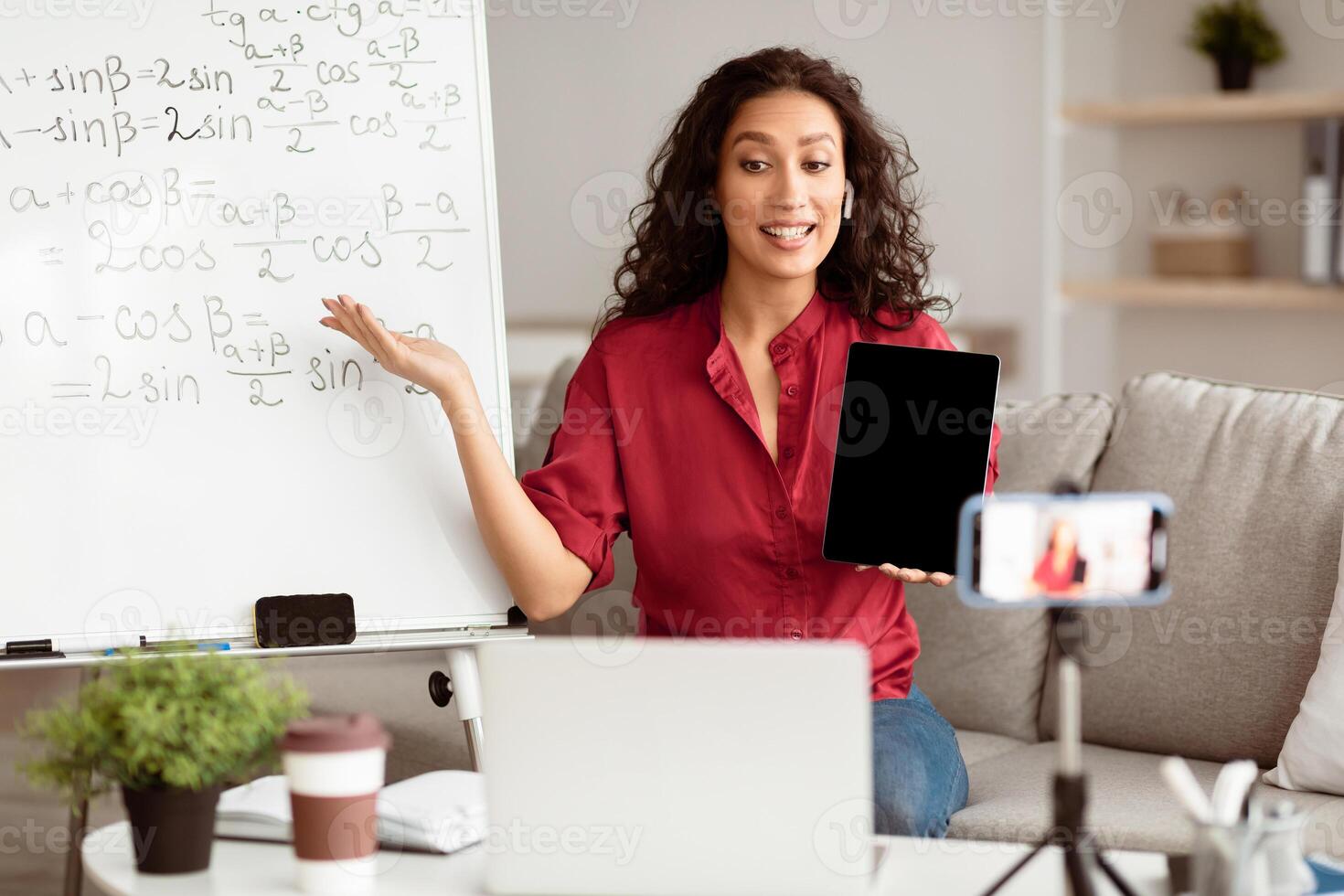 A young Middle Eastern teacher engages students through an online platform. She uses a tablet while explaining concepts in front of a whiteboard filled with equations. photo