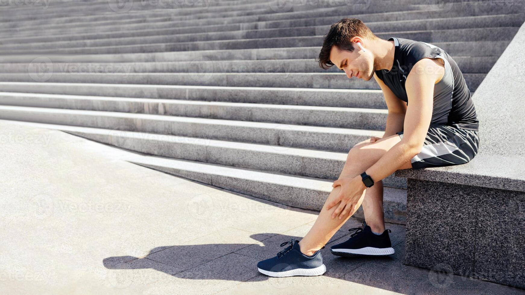 Leg injury while jogging. Guy with expression of pain on face, sits and rubs leg on street, free space photo