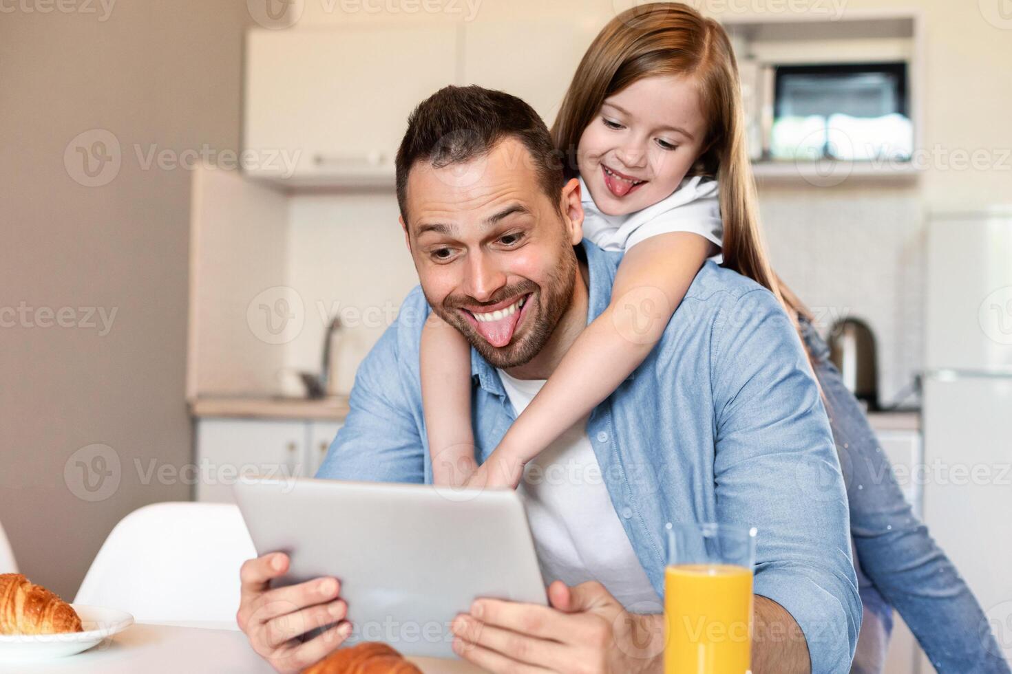 Happy Daughter Embracing Daddy Making Selfie On Digital Tablet Posing Having Fun And Showing Tongues To Computer Sitting In Kitchen At Home. Weekend Leisure, Family Bonding Using Gadgets photo